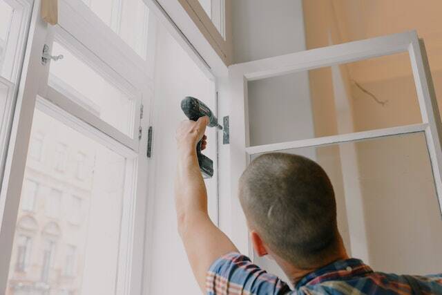 a contractor in a plaid shirt using a drill to fix a windows hinge